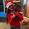 woman, dog, indoor, holiday, christmas, hat, red_clothing, glasses, kneeling, carpet, furniture, blanket, dog_bed, crocheted_blanket, smiling, long_hair, casual, portrait, festive, home