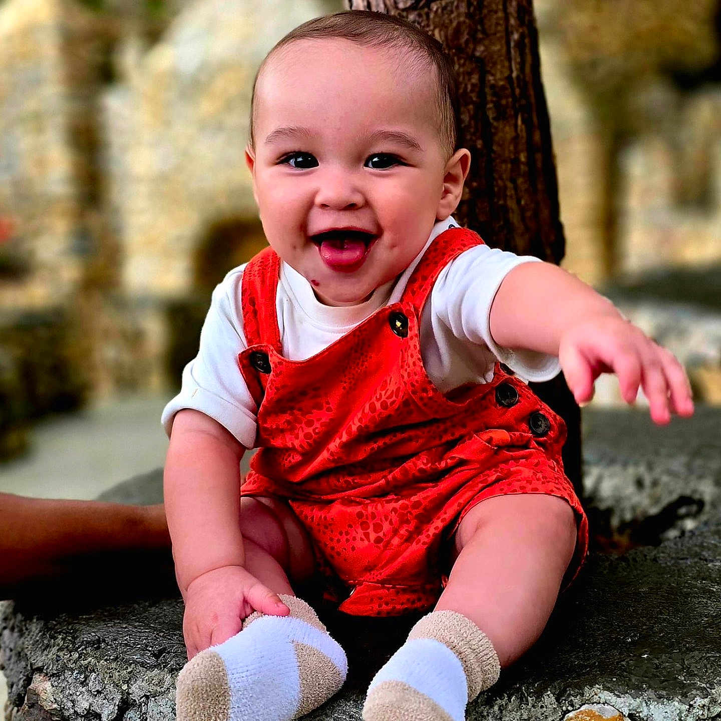 Oliver is registered to the contest to win money with this photo: baby, casual_clothing, child, cute, happy, infant, natural_light, orange_overalls, outdoor, person, playful, portrait, sitting, smiling, socks, stone, texture, toddler, tongue_out, tree_trunk