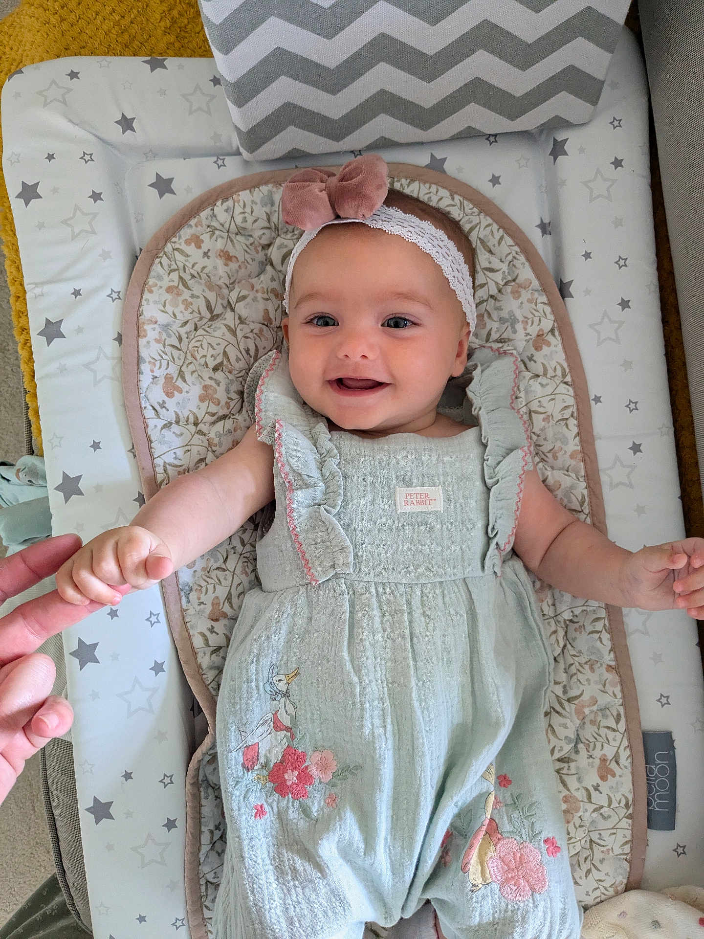 Birdy is registered to the contest to win money with this photo: baby, infant, smile, holding_hand, headband, bow, romper, floral, duck, baby_mat, pattern, star_pattern, hand, cute, happy, lying_down, indoors, person, child, soft_lighting