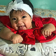 Scarlett is registered to the contest to win money with this photo: baby, headband, red_dress, smile, tongue_out, cushion, patterned_cushion, baby_clothing, infant, child, cute, happy, indoor, portrait, face, hand, lying_down, decor, bedroom, headwear