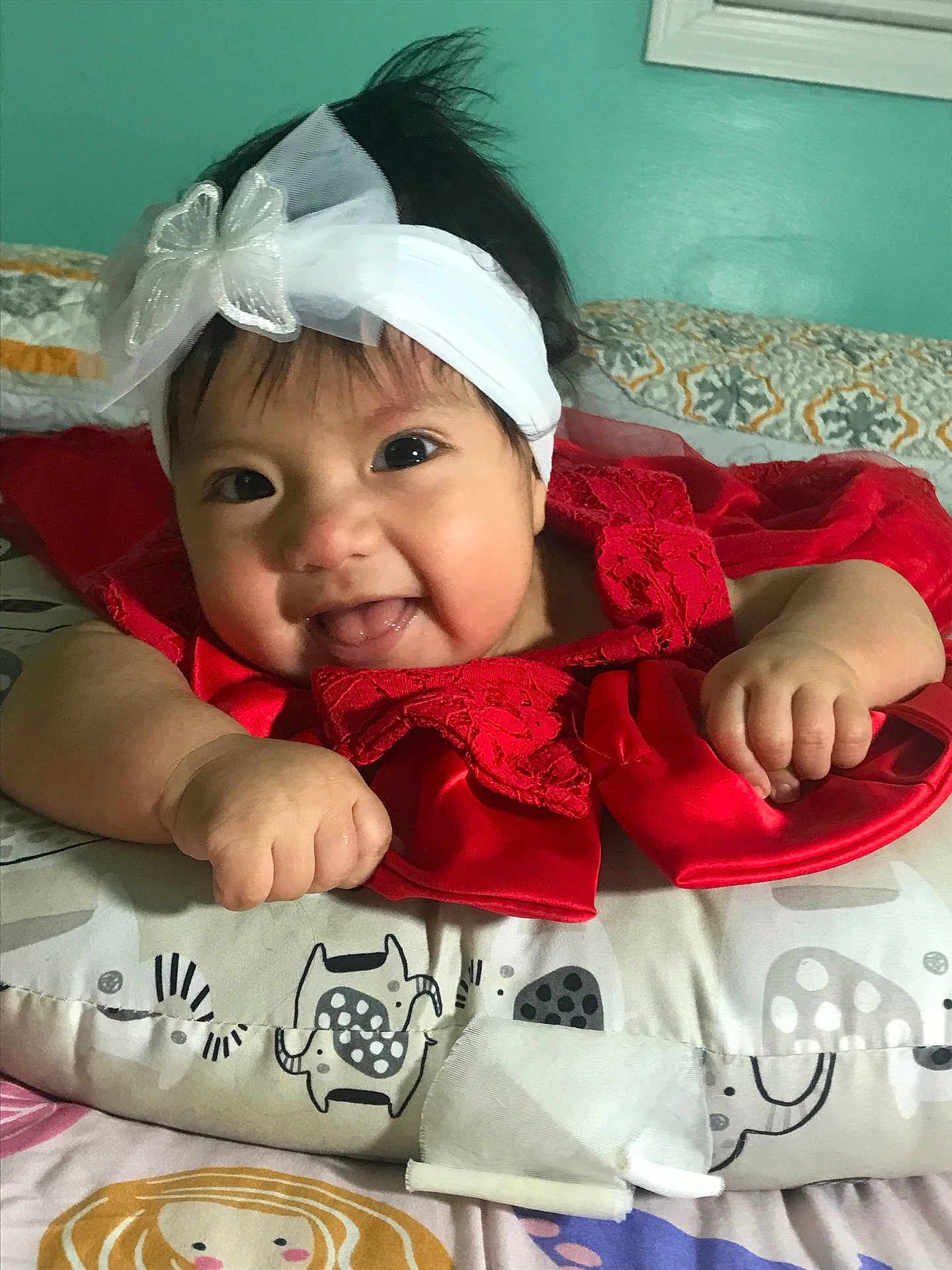 Scarlett is registered to the contest to win money with this photo: baby, headband, red_dress, smile, tongue_out, cushion, patterned_cushion, baby_clothing, infant, child, cute, happy, indoor, portrait, face, hand, lying_down, decor, bedroom, headwear