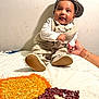 baby, child, smiling, sitting, hat, vest, bow_tie, hand, adult_hand, bracelet, henna, lentils, beans, blanket, indoors, wall, footwear, portrait, happy, clothing