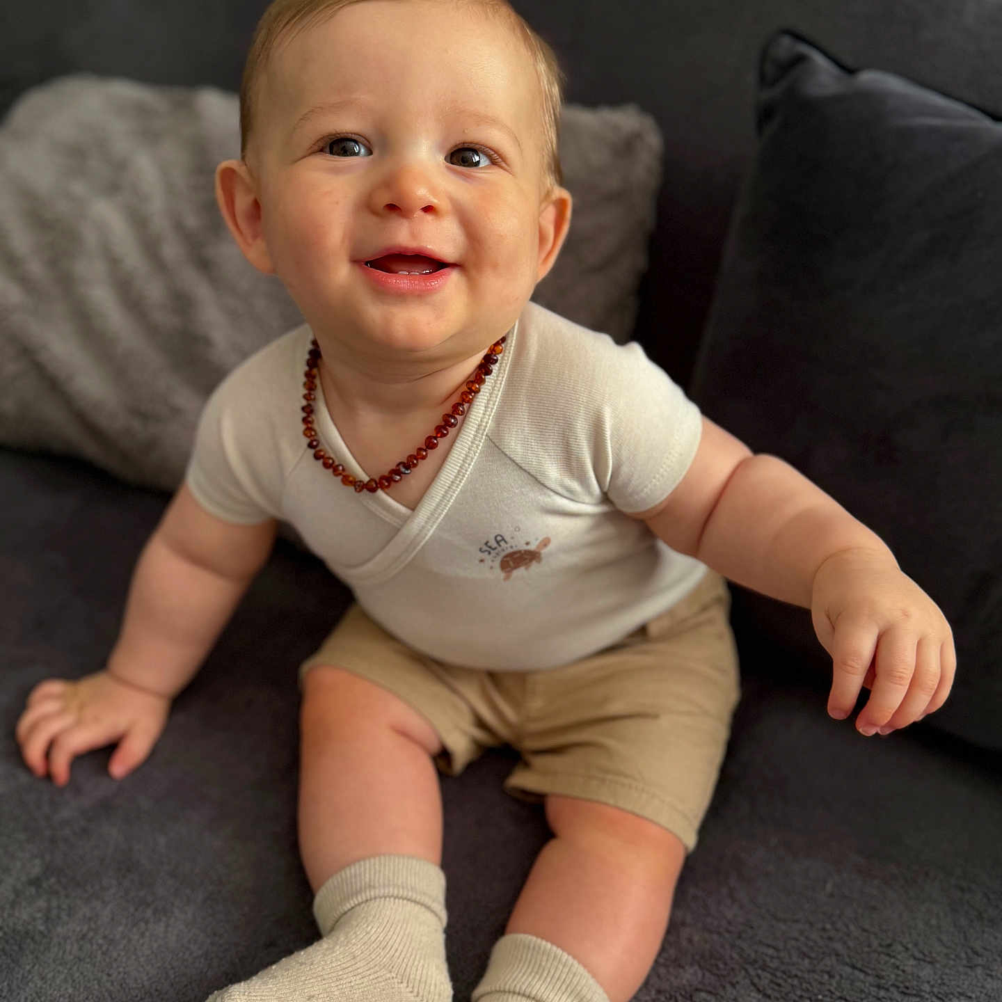 Noélio a rejoint le concours — aidez-le/la à gagner de superbes lots ! amber_necklace, baby, beige, child, clothing, couch, cute, face, happy, head, indoor, person, pillow, portrait, shorts, sitting, skin, smiling, socks, young_child