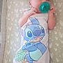 baby, cartoon_character, child, cute, fabric, hands_near_face, heart_pattern, indoors, infant, lying_down, mattress, newborn, onesie, pacifier, peaceful, sleeping_area, sleepwear, small, soft_lighting, stitch