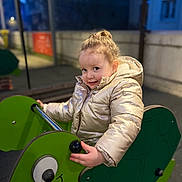 Mira participe au concours pour gagner de l'argent avec cette photo : child, smile, face, coat, puffer_jacket, playground, ride_on, green_toy, hand, hair, portrait, evening, bokeh, outdoor, urban, railing, happy, seat, toy_vehicle, curious