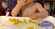 Jay Oliver is registered to the contest to win money with this photo: toddler, child, high_chair, macaroni_and_cheese, messy, food, eating, hand_to_mouth, blue_eyes, curly_hair, indoor, table, plastic_tray, snack, purple_food_pouch, face, arm, pink_chair, expression, person