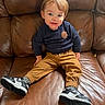 toddler, child, smiling, sitting, couch, leather, brown, sweater, navy, pants, tan, shoes, sneakers, black, white, face, person, indoor, happy, cute