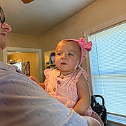 Lizzy is registered to the contest to win money with this photo: toddler, child, baby, pink_dress, headband, adult, indoor, window, blinds, light, person, holding, face, curious, smile, home, family, soft_light, casual, portrait