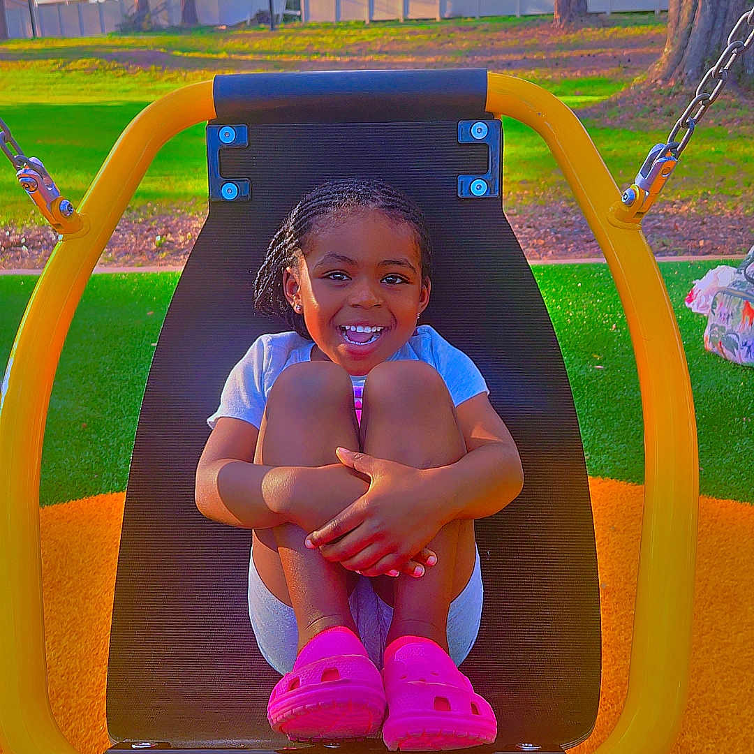 Scarlette is registered to the contest to win money with this photo: braided_hair, casual_clothing, child, daytime, fence, fun, grass, happy, nature, outdoor, park, person, pink_shoes, play, playground, seat, smiling, sunny, swing, trees