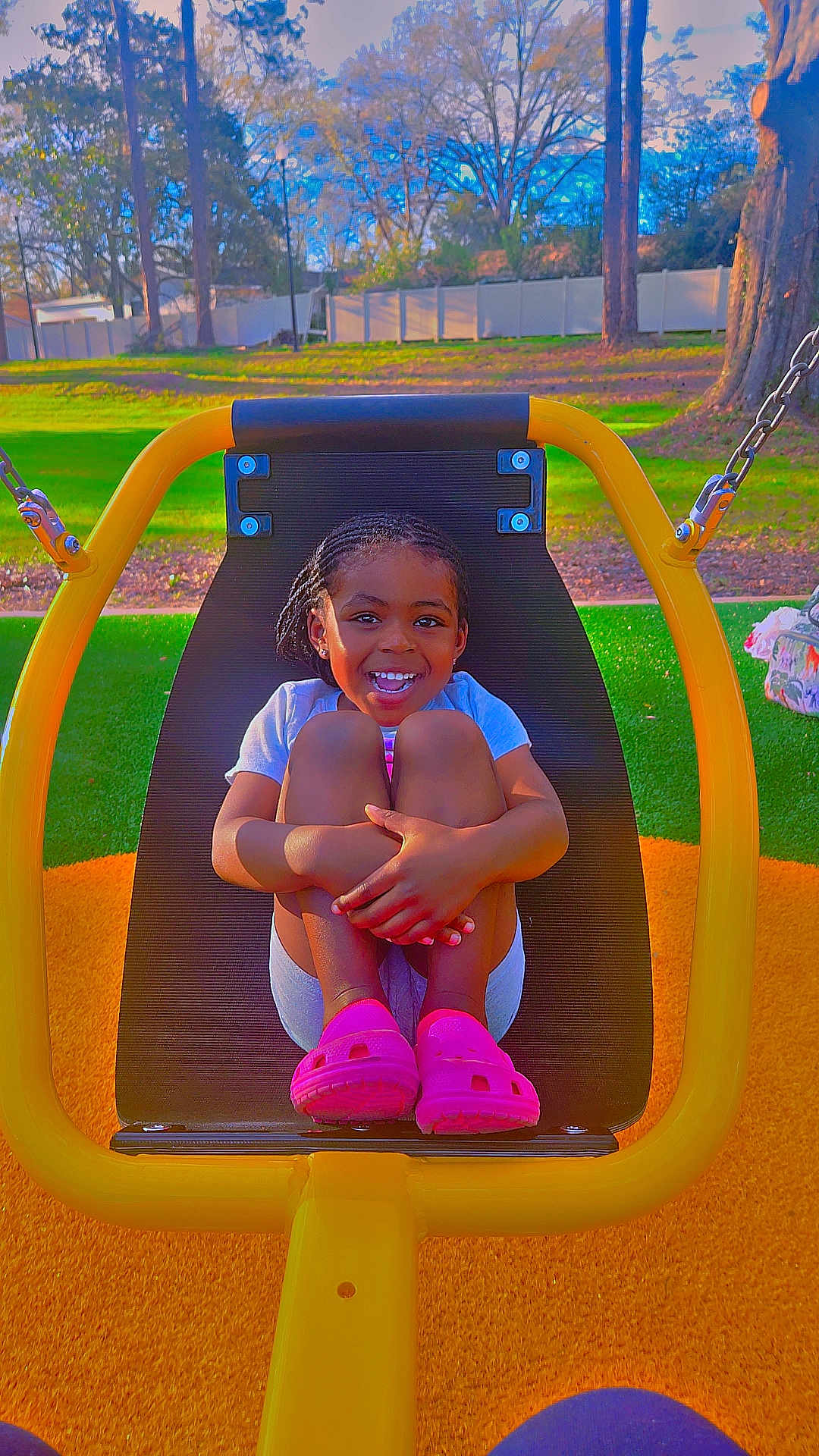 Scarlette is registered to the contest to win money with this photo: child, smiling, swing, playground, outdoor, grass, trees, braided_hair, pink_shoes, sunny, happy, seat, park, daytime, fence, casual_clothing, nature, play, person, fun