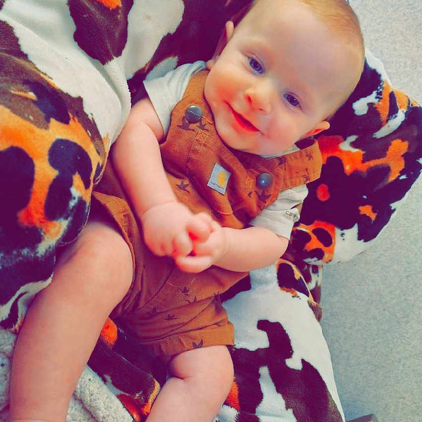 Dawson is registered to the contest to win money with this photo: arms, baby, blanket, brown_overalls, child, clothing, cozy, cute, face, happy, indoor, infant, legs, patterned_blanket, portrait, sitting, skin, smiling, soft_texture, young_child