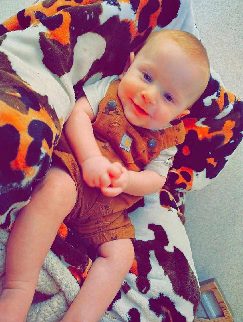 Dawson is registered to the contest to win money with this photo: baby, infant, smiling, blanket, brown_overalls, cute, cozy, child, face, skin, happy, sitting, indoor, soft_texture, patterned_blanket, legs, arms, clothing, portrait, young_child