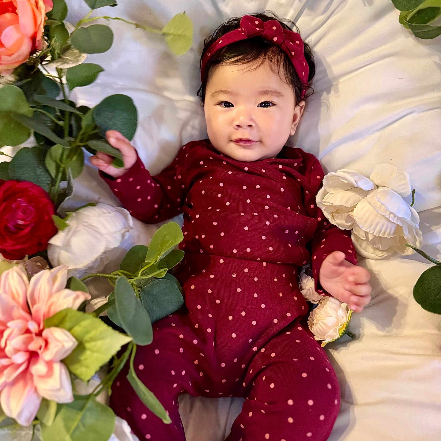 Karleen Renee joined the competition — help win amazing prizes! artificial_flowers, baby, child, closeup, cute, face, flowers, green_leaves, hand, headband, infant, lying_down, maroon_clothing, pink, polka_dot, portrait, red, smiling, soft_lighting, white_bed