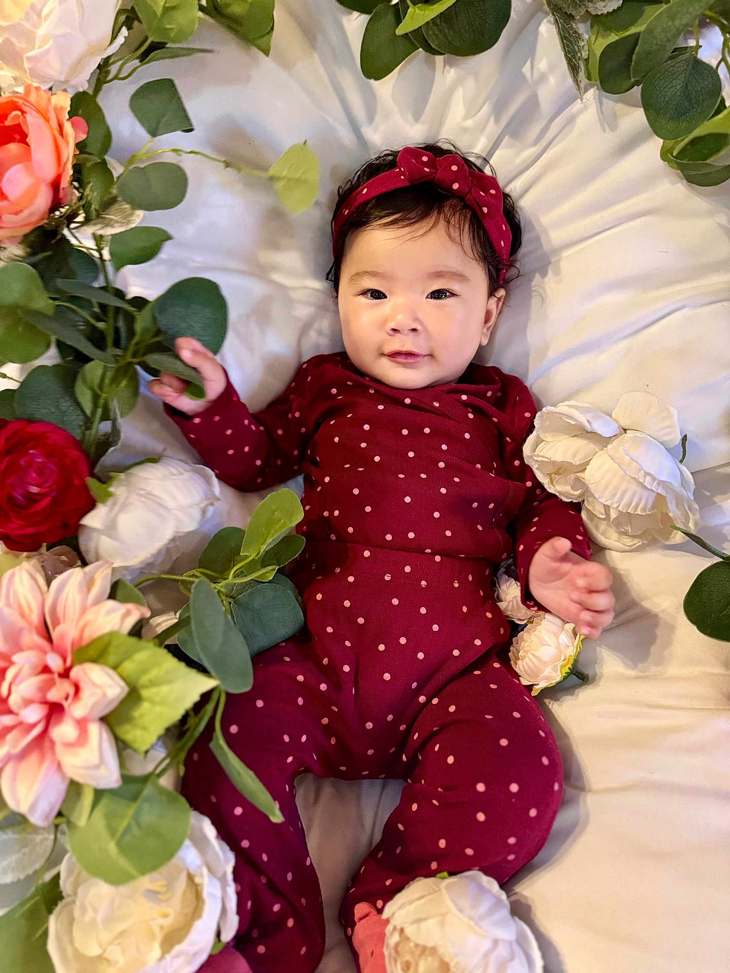 Karleen Renee joined the competition — help win amazing prizes! baby, infant, child, polka_dot, headband, maroon_clothing, flowers, artificial_flowers, green_leaves, white_bed, smiling, portrait, cute, lying_down, soft_lighting, closeup, face, hand, pink, red