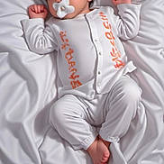 Amani Leila participe au concours pour gagner de l'argent avec cette photo : baby, infant, pacifier, white_clothing, bed, bedding, sleepwear, child, portrait, cute, newborn, relaxed, lying_down, soft_texture, indoors, peaceful, small_feet, curly_hair, orange_text, skin