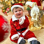Charbel is registered to the contest to win money with this photo: baby, child, smiling, santa_claus_costume, christmas_tree, holiday_decorations, festive, indoor, floor, red_clothing, white_fur_trim, sock, happy, portrait, seasonal, celebration, decor, toy, background, cute