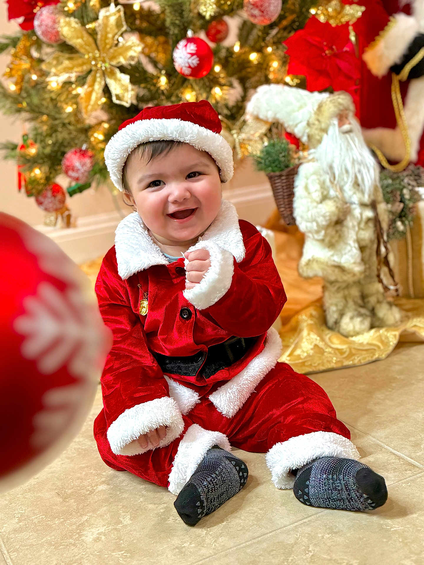 Charbel is registered to the contest to win money with this photo: baby, child, smiling, santa_claus_costume, christmas_tree, holiday_decorations, festive, indoor, floor, red_clothing, white_fur_trim, sock, happy, portrait, seasonal, celebration, decor, toy, background, cute