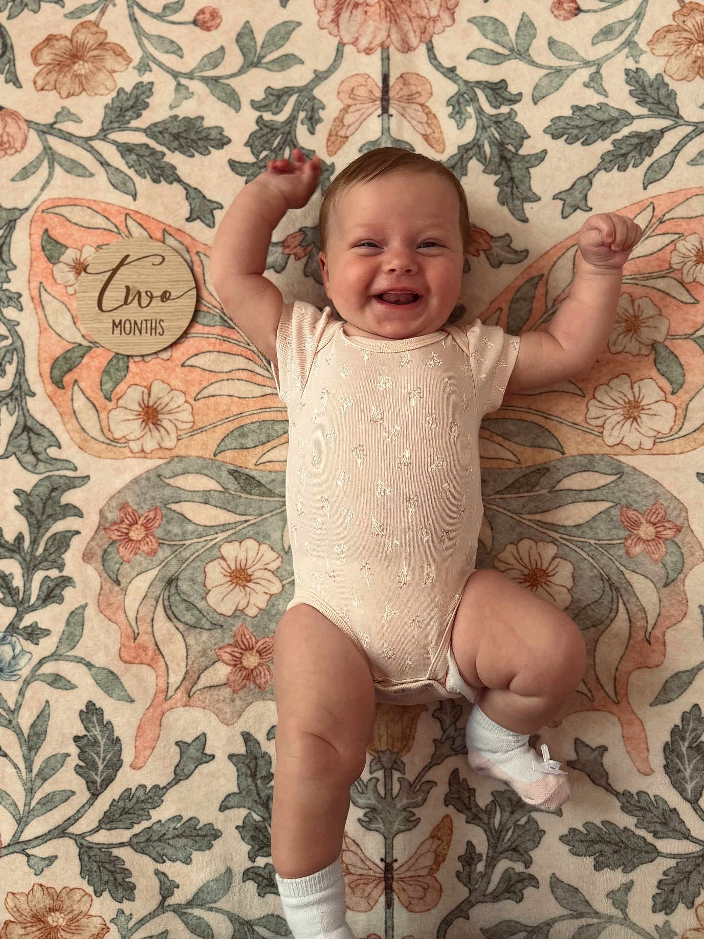 Charlotte joined the competition — help win amazing prizes! baby, smiling, onesie, white_socks, lying_down, floral_pattern, butterfly_design, wooden_plaque, two_months, milestone, happy, infant, cute, cheerful, arms_raised, blanket, floor, portrait, skin, head