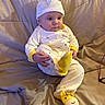 apparel, baby, child, clothing, comfort, couch, cute, face, footwear, giraffe_slippers, hat, home, indoor, infant, onesie, person, portrait, quilted_blanket, seated, soft_furniture