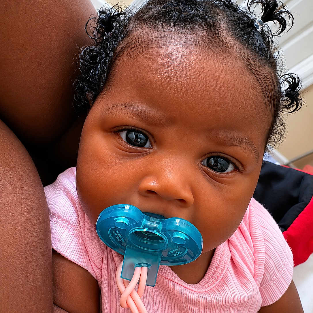 Jhené is registered to the contest to win money with this photo: baby, infant, pacifier, pink_shirt, cute, child, portrait, face, hair, curly_hair, indoor, closeup, skin, person, expression, looking_at_camera, young_child, adorable, comfort, sitting