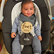 Santino is registered to the contest to win money with this photo: baby, infant, smiling, baby_rocker, blue_pajamas, plush_toys, wooden_sign, four_months, baby_slippers, indoor, child, face, happy, seated, straps, black_and_white, flooring, home, cute, portrait