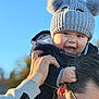 baby, child, hat, knit_hat, pom_poms, smiling, happy, person, adult, glasses, outdoor, blue_sky, hand, face, jacket, warm_clothing, portrait, family, playful, daylight