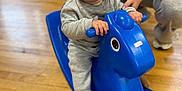 Tommy-Lee is registered to the contest to win money with this photo: baby, child, indoor, wooden_floor, rocking_horse, blue_toy, smiling, happy, person, adult_hand, clothing, socks, flooring, furniture, seat, toy, play, cute, young_child, support