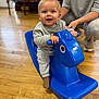 baby, child, indoor, wooden_floor, rocking_horse, blue_toy, smiling, happy, person, adult_hand, clothing, socks, flooring, furniture, seat, toy, play, cute, young_child, support