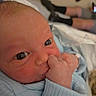 Bennett is registered to the contest to win money with this photo: newborn, baby, closeup, face, hand, thumb_sucking, blue_clothing, infant, person, skin, eyes, wrinkled_skin, bed, blanket, indoor, human, legs, socks, casual_wear, blurred_background