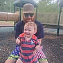 child, toddler, adult, man, beard, sunglasses, cap_hat, playground, play_structure, slide_tunnel, smile, park, outdoor, woodchips, shorts, tattoo, striped_onesie, holding, family_moment, sunlight_glare