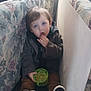 blue_shoes, brown_pants, carpet, casual_clothing, child, couch, crackers, cute, face, floral_pattern, green_container, hand, indoor, light, relaxed, sitting, snack, toddler, window, young_child