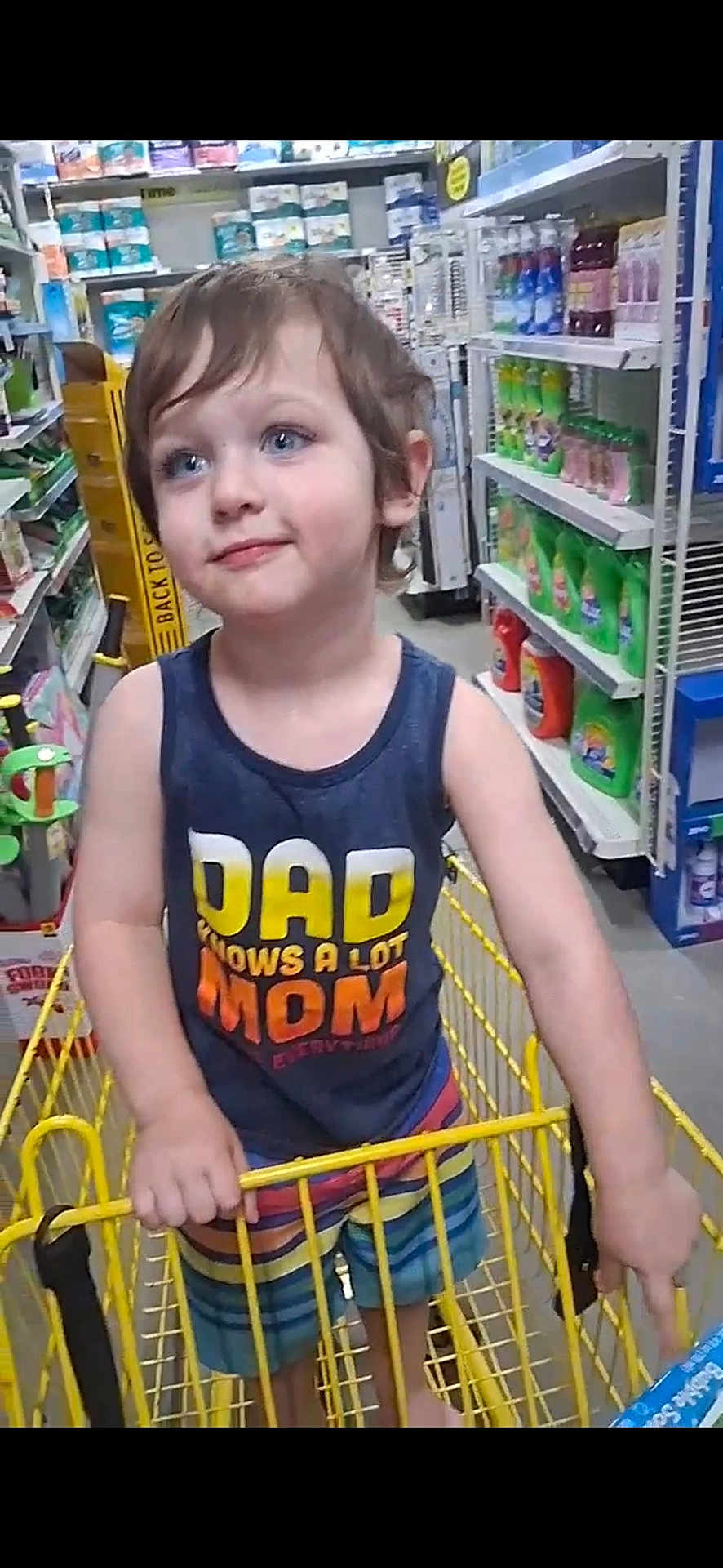 Danny is registered to the contest to win money with this photo: child, shopping_cart, store_aisle, blue_eyes, tank_top, colorful_shorts, grocery_store, household_products, smiling, young_child, tousled_hair, yellow_cart, person, retail, shelves, shopping, casual_clothing, happy, portrait, indoor