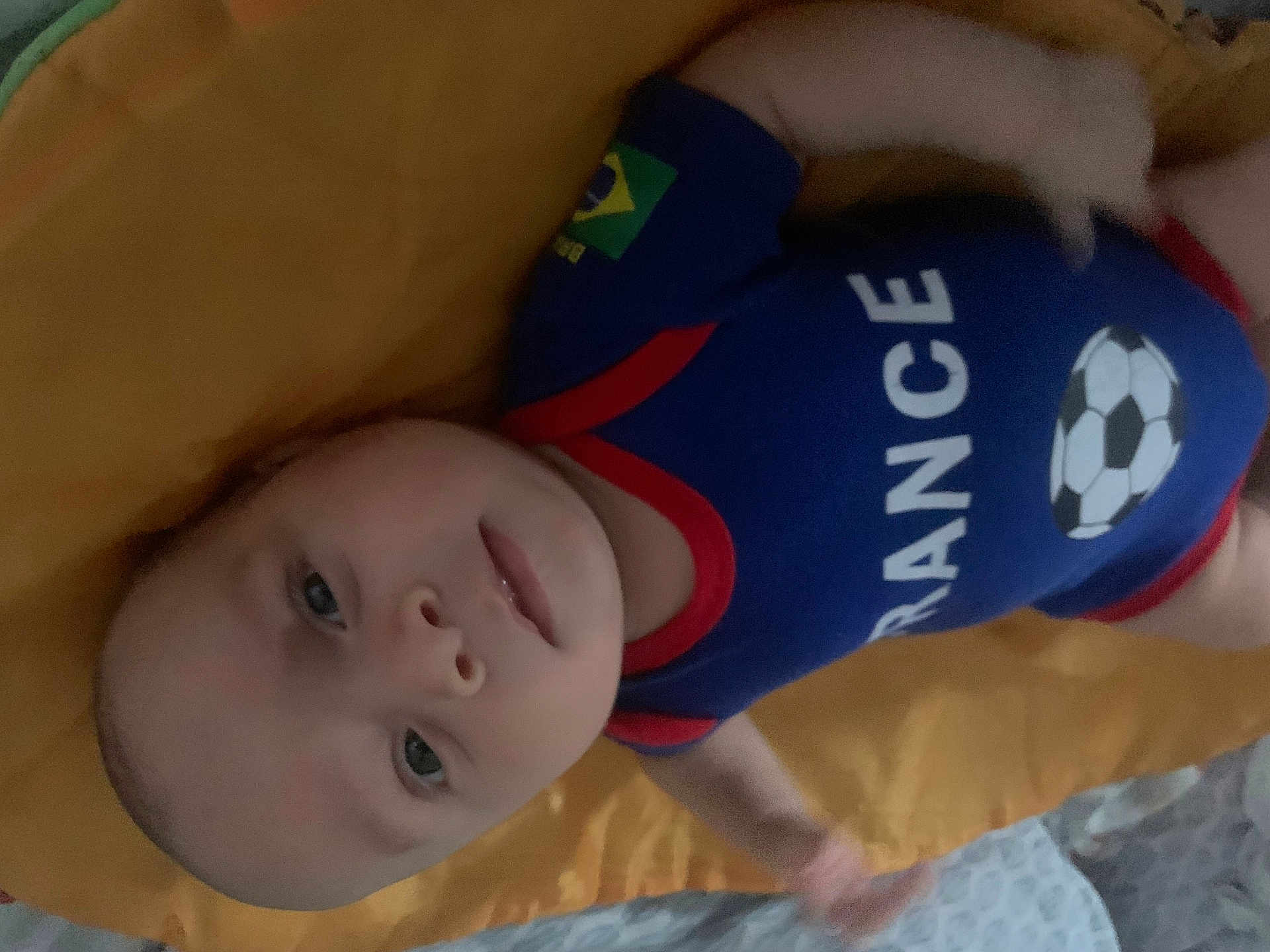 Jad participe au concours pour gagner de l'argent avec cette photo : baby, infant, blue_outfit, soccer_ball, blanket, yellow_blanket, lying_down, face, eyes, skin, person, child, cute, indoor, soft_light, clothing, head, expression, young_child, portrait