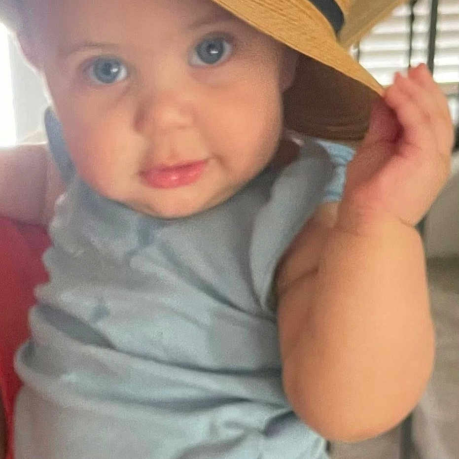 Sabina is registered to the contest to win money with this photo: baby, blue_clothing, child, closeup, curious, cute, expression, eyes, face, hand, hat, indoor, infant, person, plush_hat, portrait, sitting, smiling_hat, straw_hat, young_child