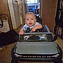 baby, toy_car, bronco, indoor, child, steering_wheel, car_toy, furniture, bookshelf, clothing, floor, door, window, room, person, car_seat, head, face, expression, hand