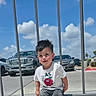 toddler, child, boy, squatting, white_tshirt, cartoon_apple, outdoor, daylight, blue_sky, clouds, metal_fence, cars, concrete, sidewalk, casual_clothing, short_hair, sneakers, playful, cute, urban