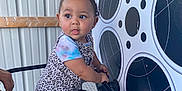 Bianca is registered to the contest to win money with this photo: toddler, child, barefoot, leopard_print, colorful_clothing, metal_chair, film_reel_graphic, indoor, curly_hair, holding, standing, railing, metal_wall, playful_expression, person, young_child, footwear_absent, looking_sideways, short_sleeves, cute