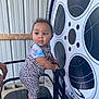 toddler, child, barefoot, leopard_print, colorful_clothing, metal_chair, film_reel_graphic, indoor, curly_hair, holding, standing, railing, metal_wall, playful_expression, person, young_child, footwear_absent, looking_sideways, short_sleeves, cute
