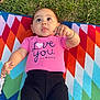 baby, child, pink_shirt, black_pants, bracelet, colorful_blanket, grass, outdoor, cute, playful, lying_down, expression, tongue_out, person, infant, casual_clothing, pattern, geometric, nature, happy
