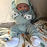 baby, infant, child, portrait, sitting, pacifier, hat, animal_hat, mittens, star_pattern, onesie, winter_clothing, socks, blanket, dinosaur_blanket, bed, indoor, cozy, looking_at_camera, baby_portrait