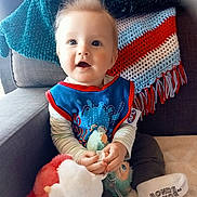 Cody is registered to the contest to win money with this photo: baby, child, plush_toy, bib, striped_clothing, couch, blanket, crocheted_throw, blue, red, white, teal, happy, smiling, indoors, person, sitting, sock, cute, soft_toy