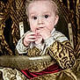 Cody is registered to the contest to win money with this photo: baby, child, infant, royal_clothing, gold, red_velvet, high_chair, ornate, patterned_fabric, curious, hands, face, portrait, indoor, luxury, decor, seated, young, expression, costume