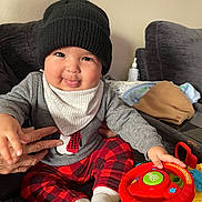 Milan is registered to the contest to win money with this photo: baby, child, toy, steering_wheel, hat, knit_hat, sweater, penguin_design, plaid_pants, red_pants, white_socks, couch, hand, blanket, indoor, playful, smiling, face, person, cozy