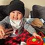 baby, child, toy, steering_wheel, hat, knit_hat, sweater, penguin_design, plaid_pants, red_pants, white_socks, couch, hand, blanket, indoor, playful, smiling, face, person, cozy