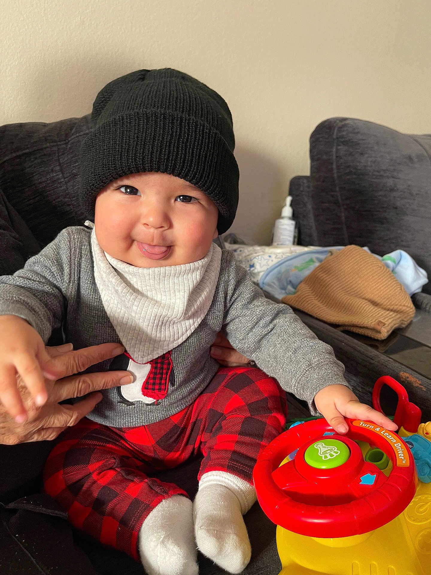Milan is registered to the contest to win money with this photo: baby, child, toy, steering_wheel, hat, knit_hat, sweater, penguin_design, plaid_pants, red_pants, white_socks, couch, hand, blanket, indoor, playful, smiling, face, person, cozy