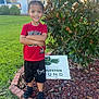 child, smile, standing, grass, tree, house, yard_sign, lawn, brick_border, red_shirt, black_shorts, sneakers, braided_hair, arms_crossed, rock_mulch, driveway, suburban, portrait, greenery, daylight