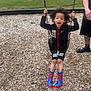 Kayden is registered to the contest to win money with this photo: child, swing, playground, wood_chips, spiderman_boots, boots, jacket, nike_logo, smiling, curly_hair, swing_chains, fence, adult_arm, sandals_on_adult, play_equipment, outdoor, grass, casual_clothing, joyful, person