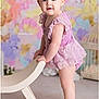baby, child, girl, standing, barefoot, chair, headband, purple_outfit, lace, pastel_background, floral, portrait, cute, smiling, indoor, person, young_child, fashion, soft_lighting, studio_photo