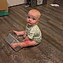 baby, child, floor, wooden_floor, plastic_container, foot, sitting, indoor, curious, looking_up, onesie, clothing, blue_eyes, light_skin, expression, home, room, door, box, flooring