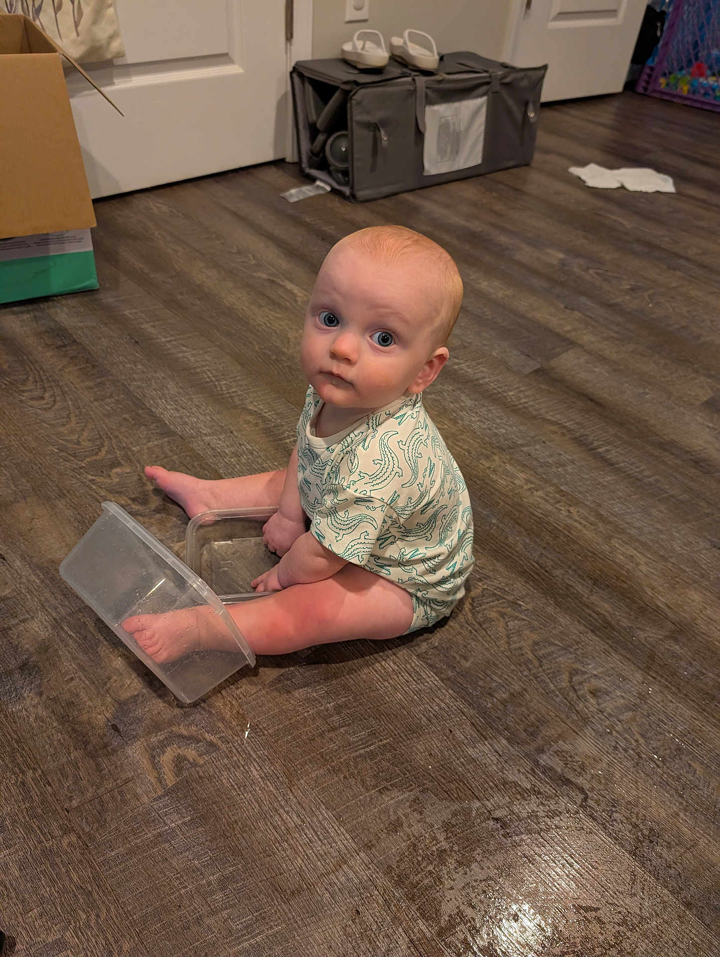 James joined the competition — help win amazing prizes! baby, child, floor, wooden_floor, plastic_container, foot, sitting, indoor, curious, looking_up, onesie, clothing, blue_eyes, light_skin, expression, home, room, door, box, flooring